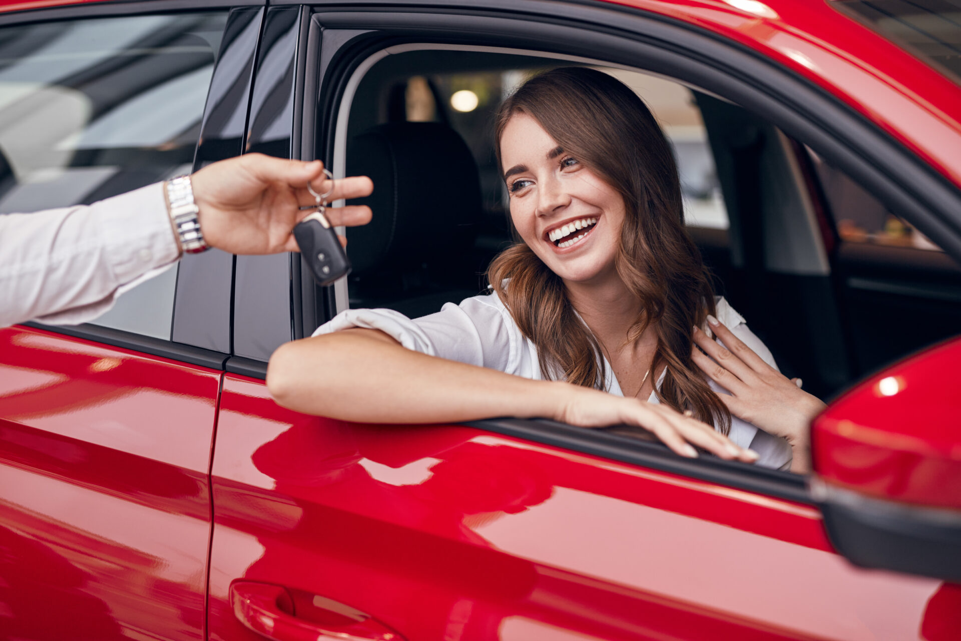 Satisfied young female sitting in new red automobile and receiving keys from unrecognizable salesman in car salon